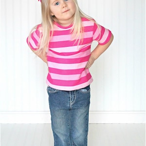 May include: A young girl wearing a pink and white striped short-sleeved shirt and blue jeans. She is standing in front of a white wall.