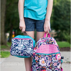 May include: A girl is carrying two bags. The larger bag is a floral patterned backpack with pink accents. The smaller bag is a lunch bag with a white and black bird pattern. The lunch bag has a turquoise zipper.