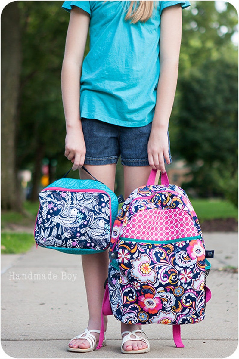 May include: A girl is carrying two bags. The larger bag is a floral patterned backpack with pink accents. The smaller bag is a lunch bag with a white and black bird pattern. The lunch bag has a turquoise zipper.