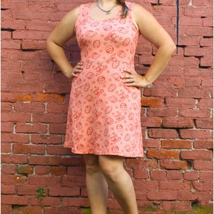 May include: A woman wearing a sleeveless peach dress with a red print of faces. The dress has a scoop neck and a fitted bodice. She is standing in front of a brick wall with green plants at her feet.