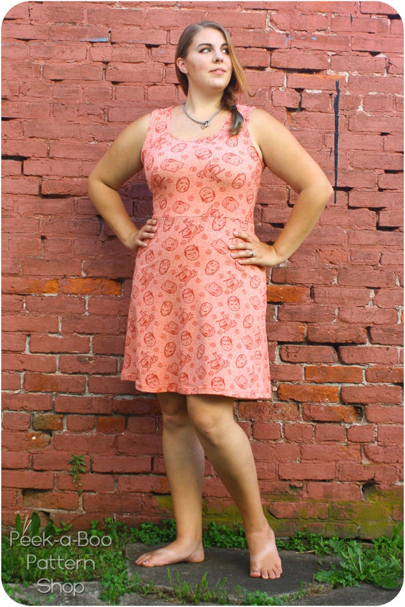 May include: A woman wearing a sleeveless peach dress with a red print of faces. The dress has a scoop neck and a fitted bodice. She is standing in front of a brick wall with green plants at her feet.