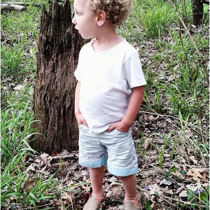 May include: A young boy with curly blonde hair wearing a white t-shirt and light blue shorts stands in a wooded area. He is looking to the left of the frame.  He is wearing orange and black sneakers.