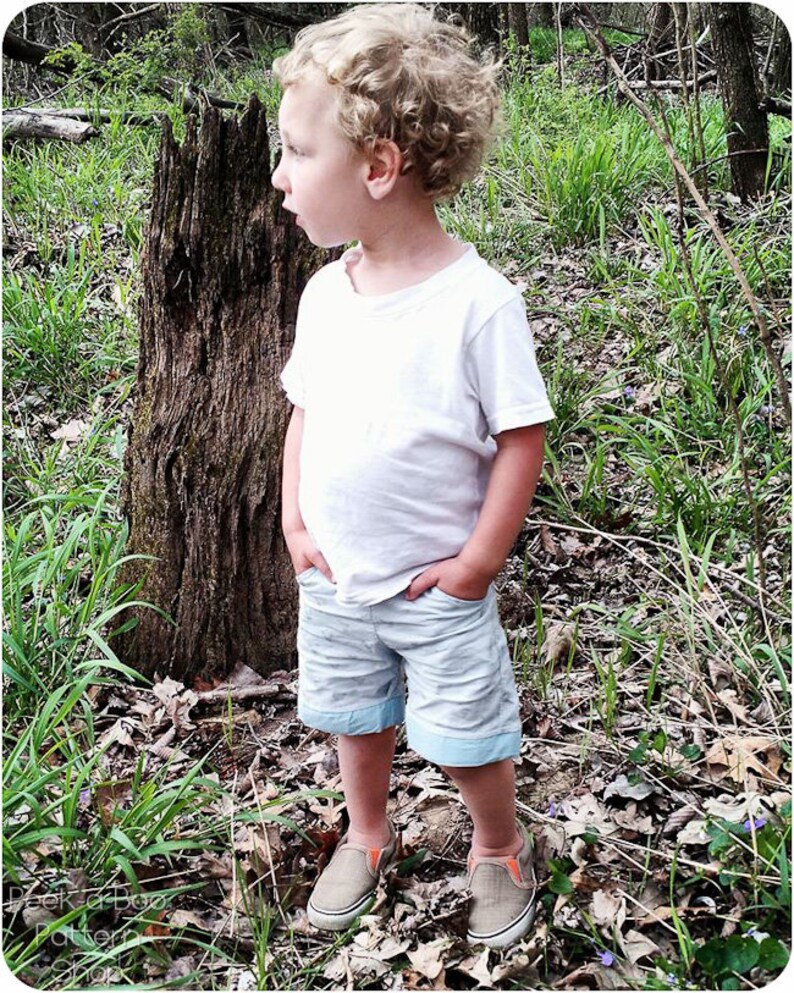 May include: A young boy with curly blonde hair wearing a white t-shirt and light blue shorts stands in a wooded area. He is looking to the left of the frame.  He is wearing orange and black sneakers.
