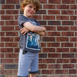 May include: A young boy with curly hair is wearing a grey t-shirt with a Star Wars graphic and grey shorts with black trim. He is standing in front of a brick wall.