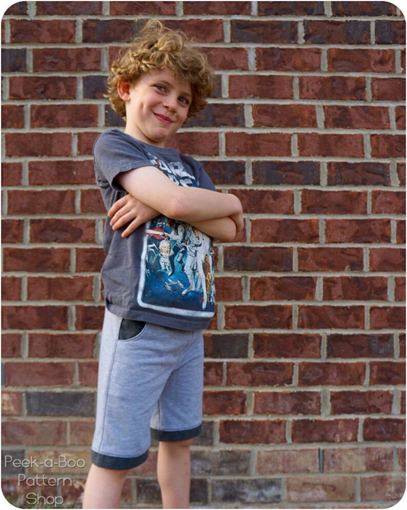 May include: A young boy with curly hair is wearing a grey t-shirt with a Star Wars graphic and grey shorts with black trim. He is standing in front of a brick wall.