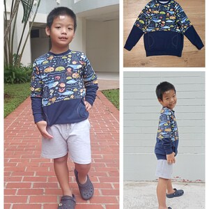 May include: A young boy wearing a blue long-sleeved sweatshirt with a colorful sea creature print and a navy blue pocket. The sweatshirt has a contrasting navy blue bottom half. He is wearing grey shorts and black slip-on shoes.