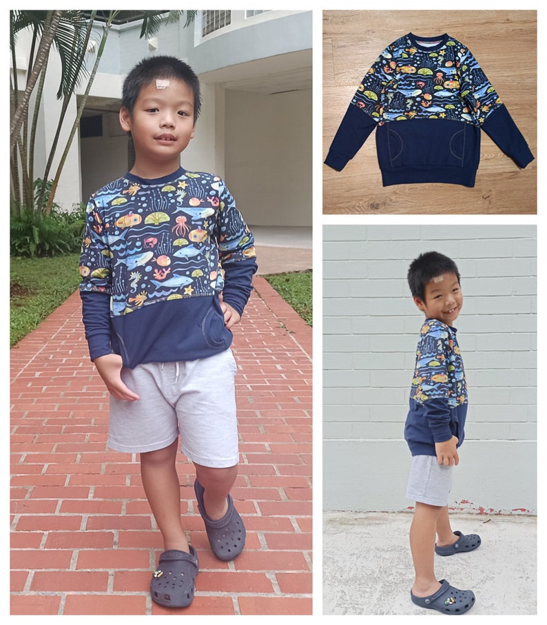 May include: A young boy wearing a blue long-sleeved sweatshirt with a colorful sea creature print and a navy blue pocket. The sweatshirt has a contrasting navy blue bottom half. He is wearing grey shorts and black slip-on shoes.