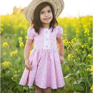 May include: A young girl wearing a pink floral dress with white buttons and a white lace trim. She is wearing a straw hat and white sandals. She is standing in a field of yellow flowers.