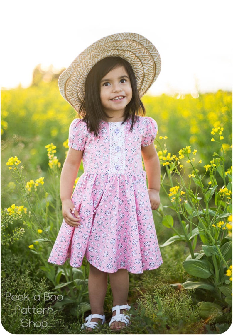 May include: A young girl wearing a pink floral dress with white buttons and a white lace trim. She is wearing a straw hat and white sandals. She is standing in a field of yellow flowers.