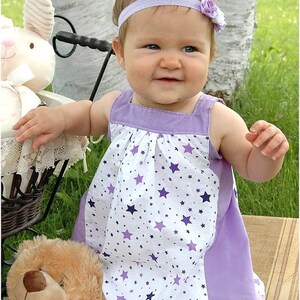 May include: A young girl wearing a white and purple dress with a star pattern, a purple headband with a flower, and white tights. She is standing next to a stuffed animal and a wicker basket.
