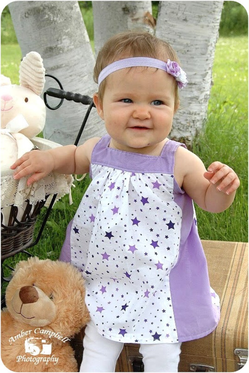 May include: A young girl wearing a white and purple dress with a star pattern, a purple headband with a flower, and white tights. She is standing next to a stuffed animal and a wicker basket.