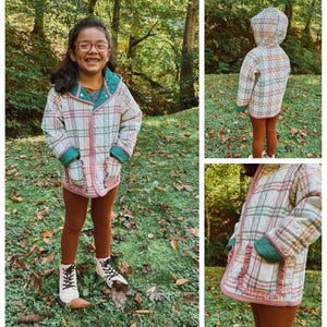May include: A child wearing a plaid quilted jacket with a green hood and brown leggings. The jacket has a ruffled trim and a pocket on each side. The child is standing in a grassy area with autumn leaves on the ground.