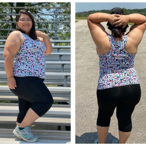 May include: A woman wearing a white tank top with a colorful geometric pattern and black capri leggings. She is standing in front of a set of bleachers.