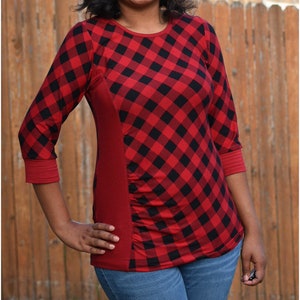 May include: A woman wearing a red and black plaid shirt with a red side panel and blue jeans. The shirt has a diamond pattern.
