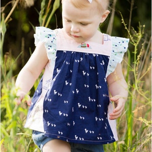 May include: A young girl wearing a blue and white patterned dress with a pink bodice and white ruffle sleeves. She is standing in a field of tall grass.