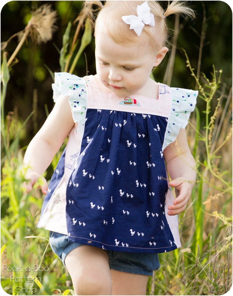 May include: A young girl wearing a blue and white patterned dress with a pink bodice and white ruffle sleeves. She is standing in a field of tall grass.