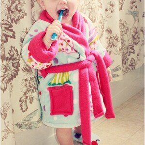 May include: A young child wearing a pink and blue fleece robe with a colorful cupcake pattern. The child is brushing their teeth with a blue toothbrush.