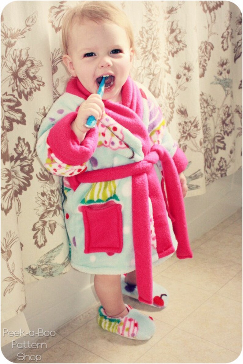 May include: A young child wearing a pink and blue fleece robe with a colorful cupcake pattern. The child is brushing their teeth with a blue toothbrush.