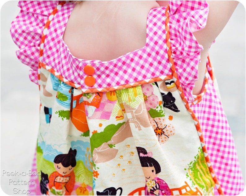 May include: A pink and white gingham dress with orange trim and a white bodice featuring a colorful floral print. The dress has a gathered skirt and a ruffled neckline.