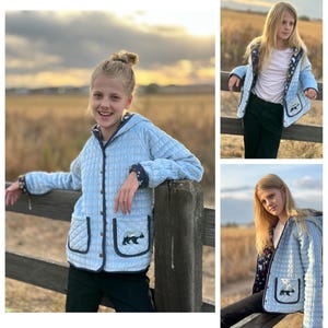 May include: A light blue quilted jacket with a hood and a bear design on the pocket. The jacket has a button closure and navy blue trim.