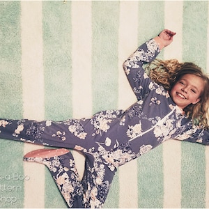 May include: A young girl wearing a purple floral print pajamas with a zipper in the front, lying on a white and green striped rug. She is smiling and looking at the camera.