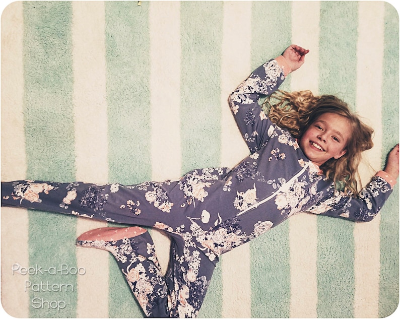 May include: A young girl wearing a purple floral print pajamas with a zipper in the front, lying on a white and green striped rug. She is smiling and looking at the camera.