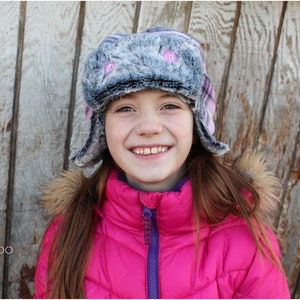 May include: A young person wearing a pink puffy jacket with a purple zipper and a gray and pink faux fur hat with pink buttons. The hat has ear flaps that are folded down.