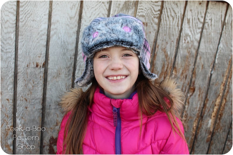 May include: A young person wearing a pink puffy jacket with a purple zipper and a gray and pink faux fur hat with pink buttons. The hat has ear flaps that are folded down.