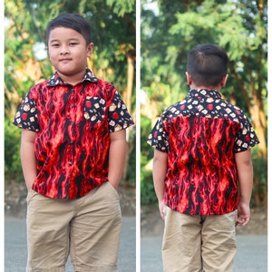 Puede incluir: Una persona joven lleva una camisa de manga corta con botones de color rojo y negro con un patr&oacute;n de llamas y un fondo blanco. La camisa tiene una variedad de peque&ntilde;as im&aacute;genes de alimentos. La persona tambi&eacute;n lleva pantalones cortos de color caqui.
