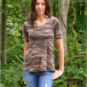 May include: A woman wearing a brown and green camouflage print top with short sleeves and blue jeans with distressed details. The top has a scoop neckline and a slightly flared hem.