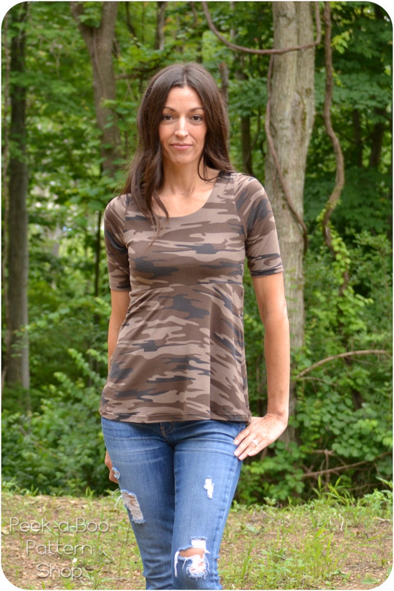 May include: A woman wearing a brown and green camouflage print top with short sleeves and blue jeans with distressed details. The top has a scoop neckline and a slightly flared hem.