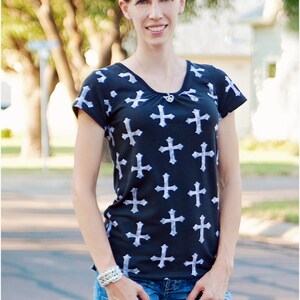 May include: A woman wearing a black and white t-shirt with a cross pattern and blue jeans. She is standing outdoors with trees and a building in the background.