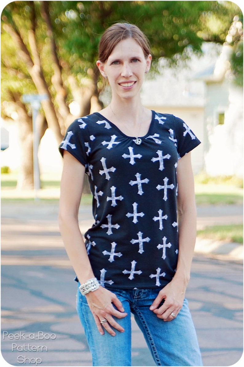 May include: A woman wearing a black and white t-shirt with a cross pattern and blue jeans. She is standing outdoors with trees and a building in the background.