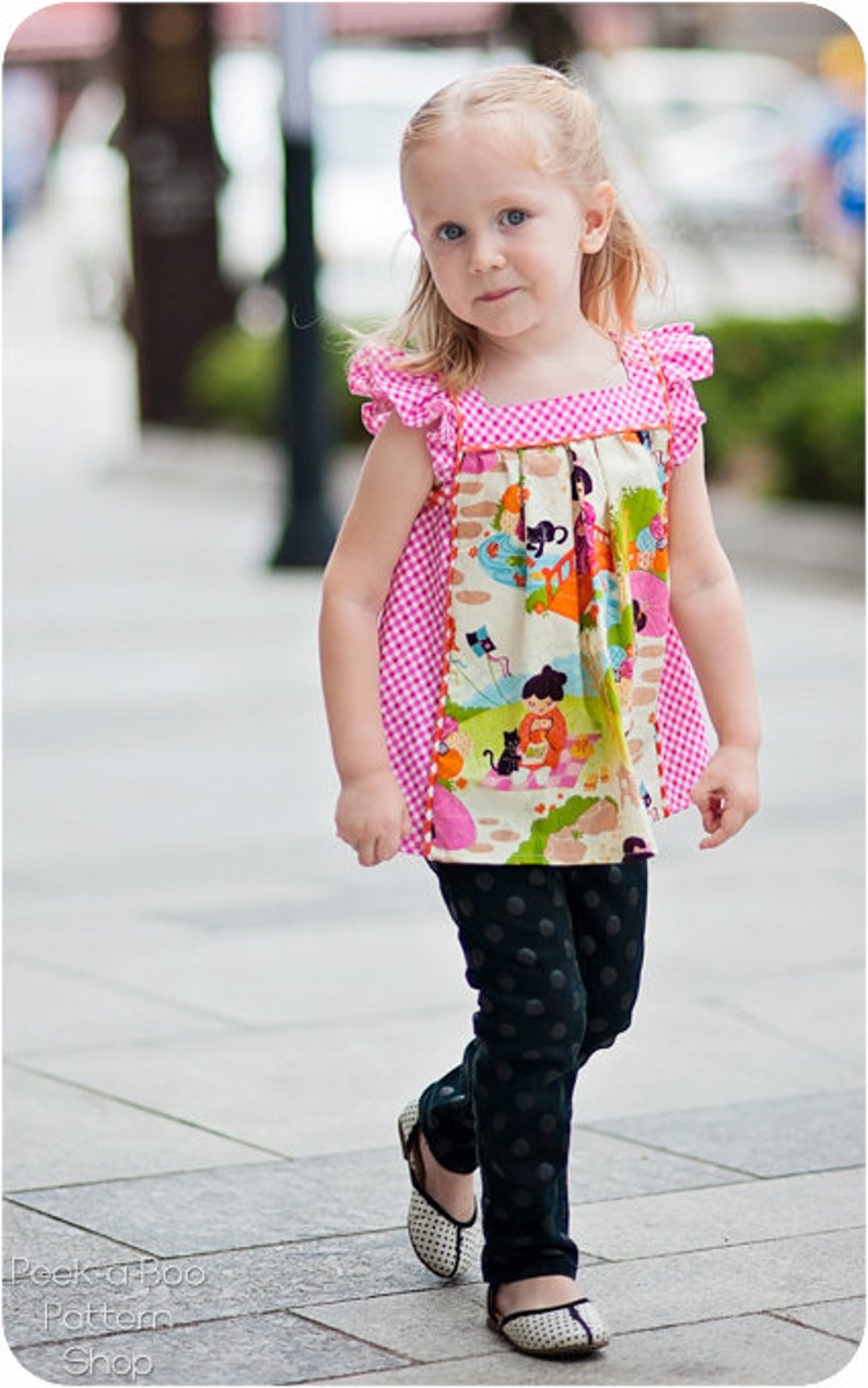 May include: A young girl wearing a pink and white gingham top with a colorful print and black polka dot pants. She is wearing white shoes with black accents.