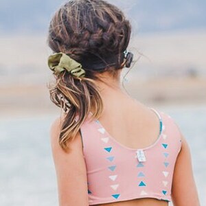 May include: A young person with long brown hair wearing a pink and blue patterned swimsuit top. The top has a scoop neck and a racerback style. The person is facing away from the camera and looking at something in the distance.