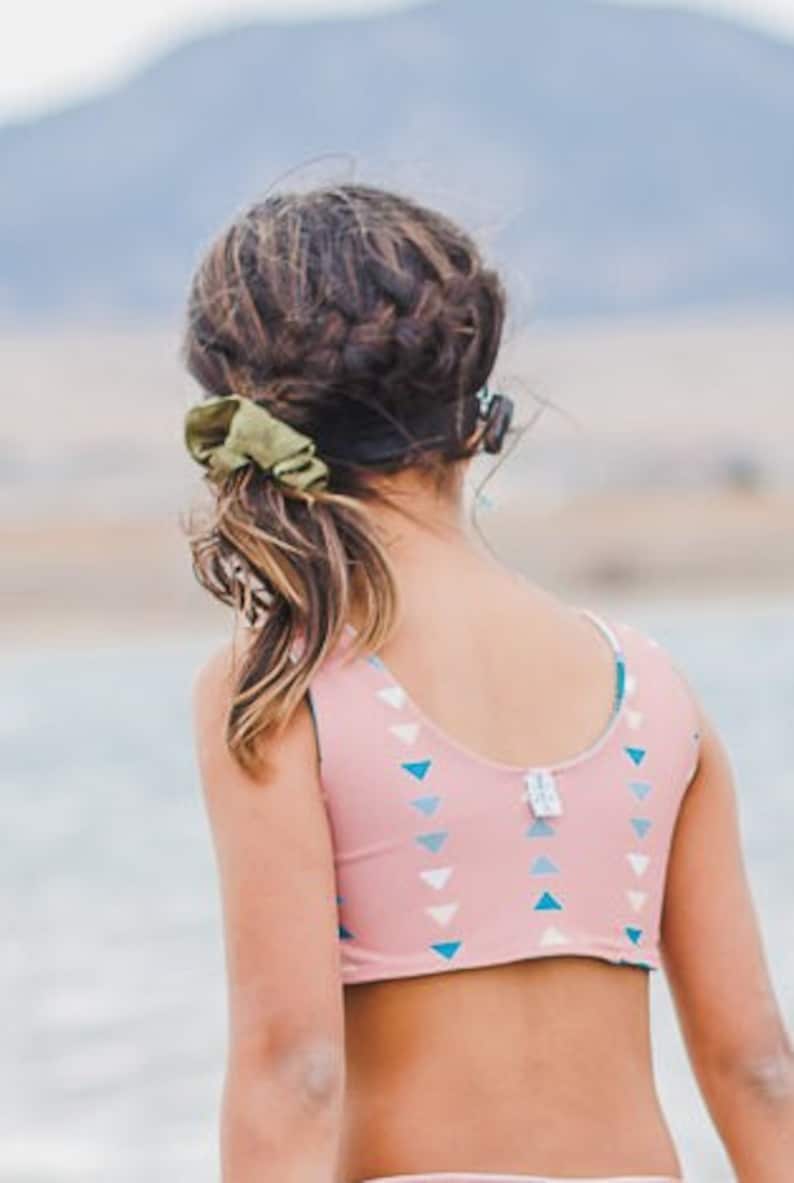 May include: A young person with long brown hair wearing a pink and blue patterned swimsuit top. The top has a scoop neck and a racerback style. The person is facing away from the camera and looking at something in the distance.