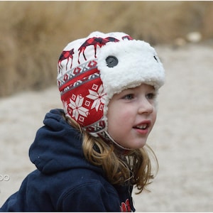 May include: A child wearing a red and white patterned winter hat with a white faux fur ear flap. The hat has a moose design and a snowflake pattern.