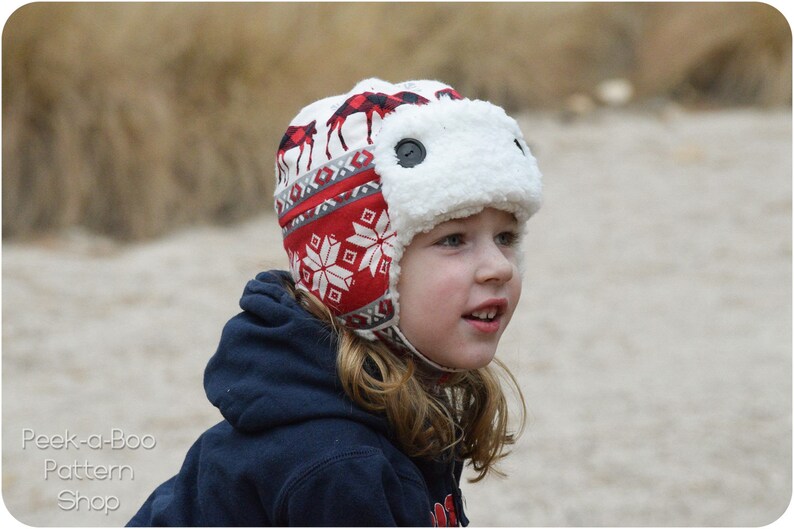 May include: A child wearing a red and white patterned winter hat with a white faux fur ear flap. The hat has a moose design and a snowflake pattern.