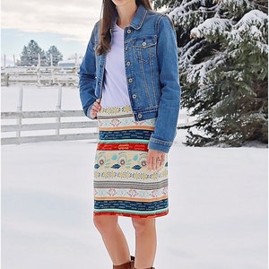 May include: A woman wearing a white t-shirt, a blue denim jacket, and a colorful patterned skirt stands in a snowy field. She is wearing brown ankle boots. The skirt has a geometric pattern with red, blue, yellow, and white colors. The text "Peek-a-Boo Pattern Shop" is visible in the lower left corner of the image.