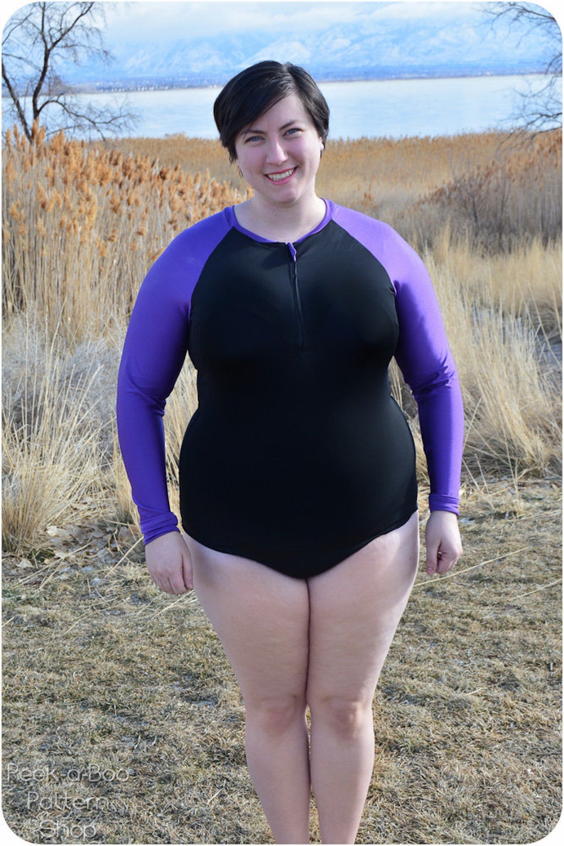 May include: A woman wearing a black and purple long-sleeved swimsuit with a zipper in the front. She is standing in a field of tall grass with a body of water in the background.