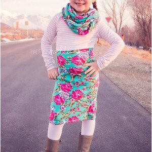 Puede incluir: Una ni&ntilde;a peque&ntilde;a con una camisa de manga larga a rayas blancas y rosas, una bufanda infinita floral de color turquesa y rosa, una falda floral de color turquesa y rosa, medias blancas y botas marrones. La ni&ntilde;a est&aacute; de pie en un camino pavimentado con una cordillera de monta&ntilde;as en el fondo. El texto "MADE BY MELLI" es visible en la esquina inferior izquierda de la imagen.