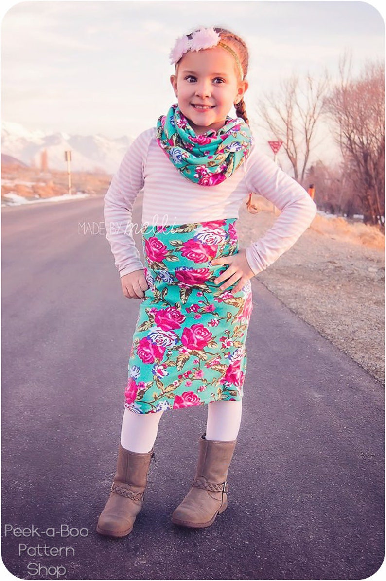 Puede incluir: Una ni&ntilde;a peque&ntilde;a con una camisa de manga larga a rayas blancas y rosas, una bufanda infinita floral de color turquesa y rosa, una falda floral de color turquesa y rosa, medias blancas y botas marrones. La ni&ntilde;a est&aacute; de pie en un camino pavimentado con una cordillera de monta&ntilde;as en el fondo. El texto "MADE BY MELLI" es visible en la esquina inferior izquierda de la imagen.