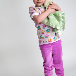 Puede incluir: Una ni&ntilde;a peque&ntilde;a lleva una camisa de manga corta rosa y blanca con un estampado de b&uacute;hos coloridos y pantalones de terciopelo morado. Sostiene una manta verde y blanca con un animal de peluche.