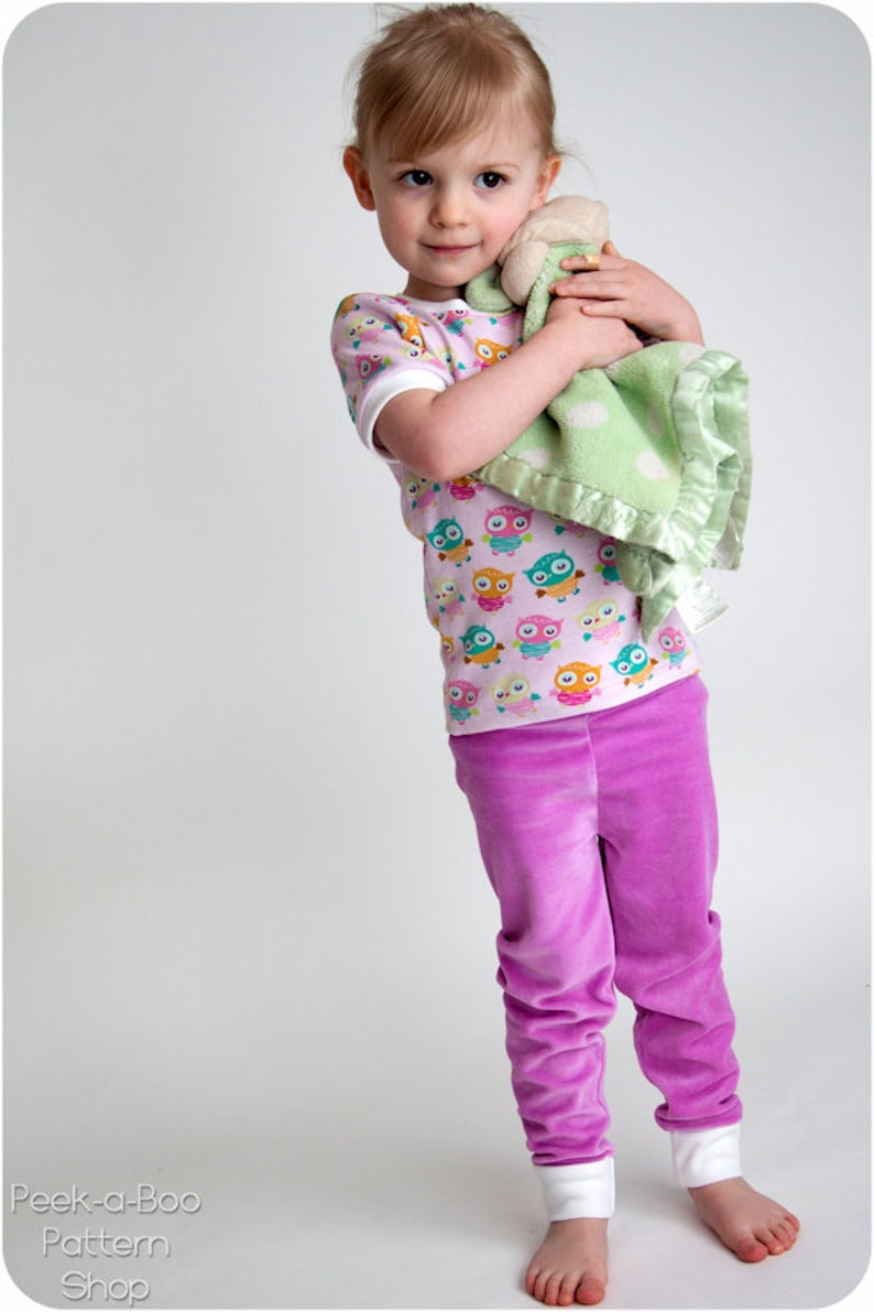 Puede incluir: Una ni&ntilde;a peque&ntilde;a lleva una camisa de manga corta rosa y blanca con un estampado de b&uacute;hos coloridos y pantalones de terciopelo morado. Sostiene una manta verde y blanca con un animal de peluche.