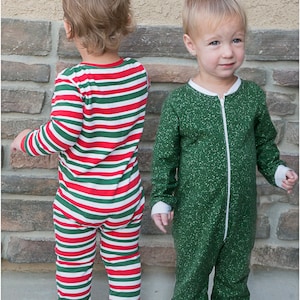 May include: Two toddlers wearing matching pajamas. One child is wearing red and white striped pajamas, and the other is wearing green pajamas with white speckles. Both pajamas have long sleeves and feet.
