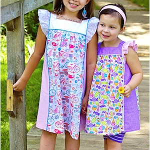 May include: Two young girls wearing colorful dresses with floral and candy patterns. The girl on the left is wearing a pink and white dress with a blue trim and a floral pattern. The girl on the right is wearing a purple and white dress with a candy pattern. Both girls are wearing sandals.