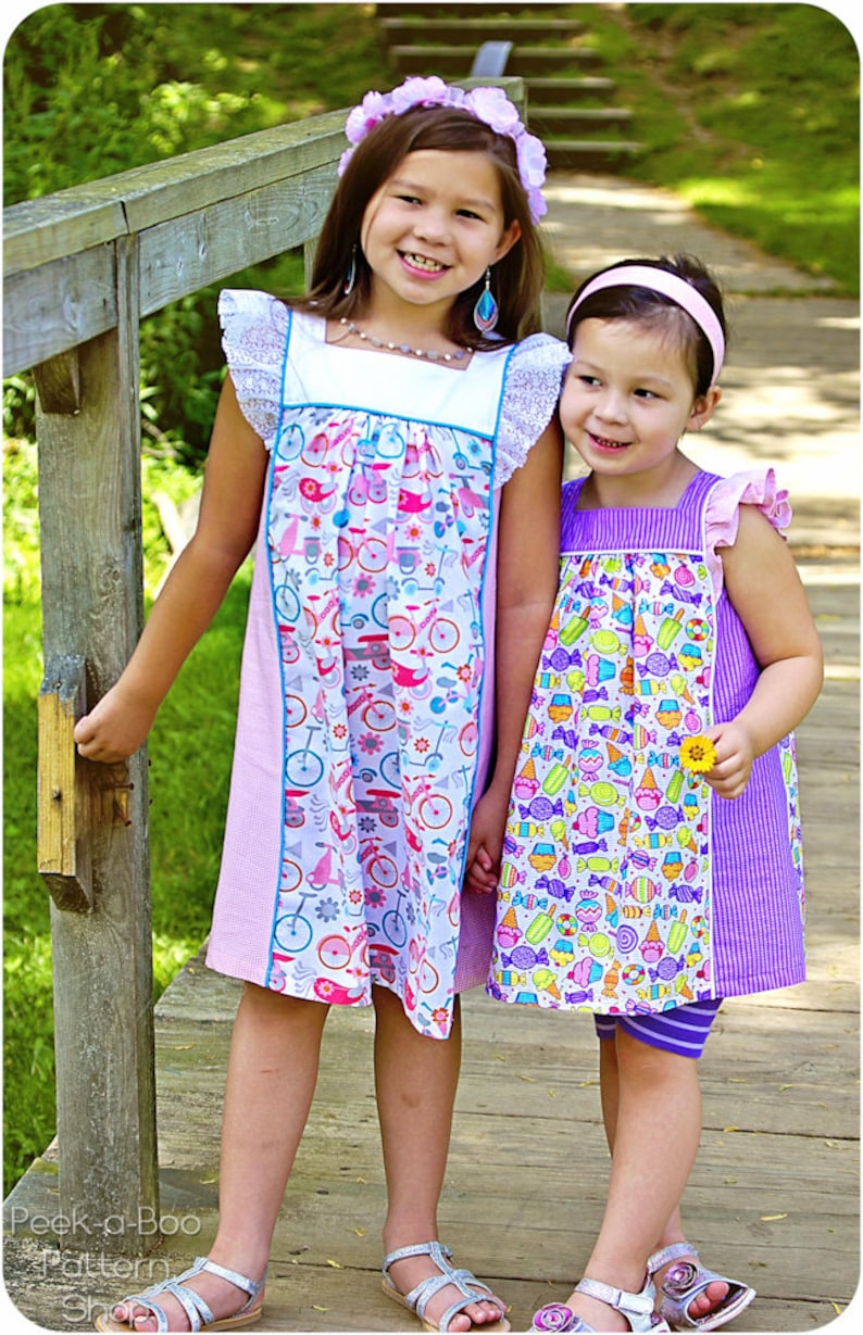 May include: Two young girls wearing colorful dresses with floral and candy patterns. The girl on the left is wearing a pink and white dress with a blue trim and a floral pattern. The girl on the right is wearing a purple and white dress with a candy pattern. Both girls are wearing sandals.