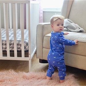 May include: A baby wearing a blue long-sleeved onesie with a white bow pattern is standing on a pink fuzzy rug. The baby is looking over their shoulder and has a gold bow in their hair.