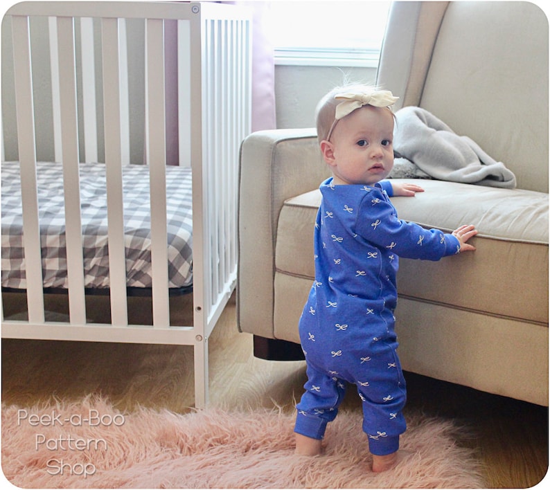 May include: A baby wearing a blue long-sleeved onesie with a white bow pattern is standing on a pink fuzzy rug. The baby is looking over their shoulder and has a gold bow in their hair.