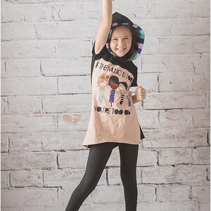 May include: A young person wearing a black and pink striped sun hat, a beige t-shirt with a graphic of four people and the text "If the music is too loud you're too old", and black leggings. The person is standing in front of a white brick wall.